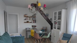 Ein Wohnzimmer mit einem petrolfarbenen Sofa, einem Essbereich mit einem Glastisch und bunten Stühlen, einem weißen Schrank und einer Treppe mit dunklem Holzbelag.
