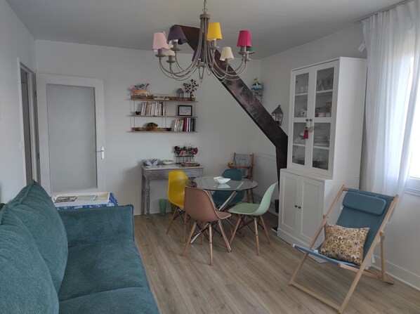 A living room with a teal sofa, a dining area with a glass table and colorful chairs, a white cabinet, and a staircase with a dark wood finish.