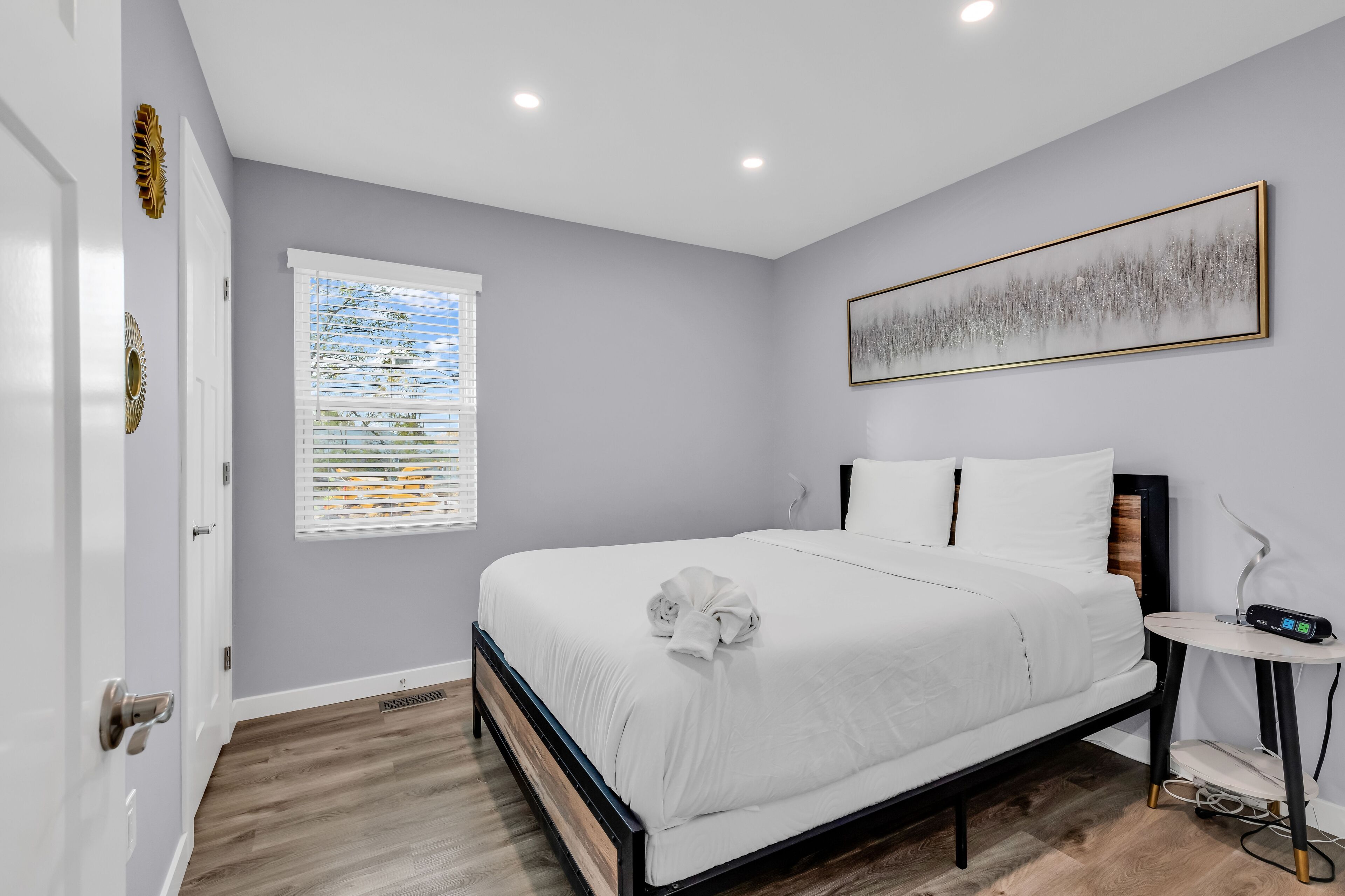 Une chambre à coucher comprenant un lit, une table de chevet, une fenêtre avec des stores et une œuvre d’art accrochée au mur.
