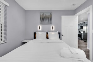 Moderni hotellihuone, jossa on suuri sänky, yöpöydät, taulu seinällä ja näkymä keittiötilaan.
