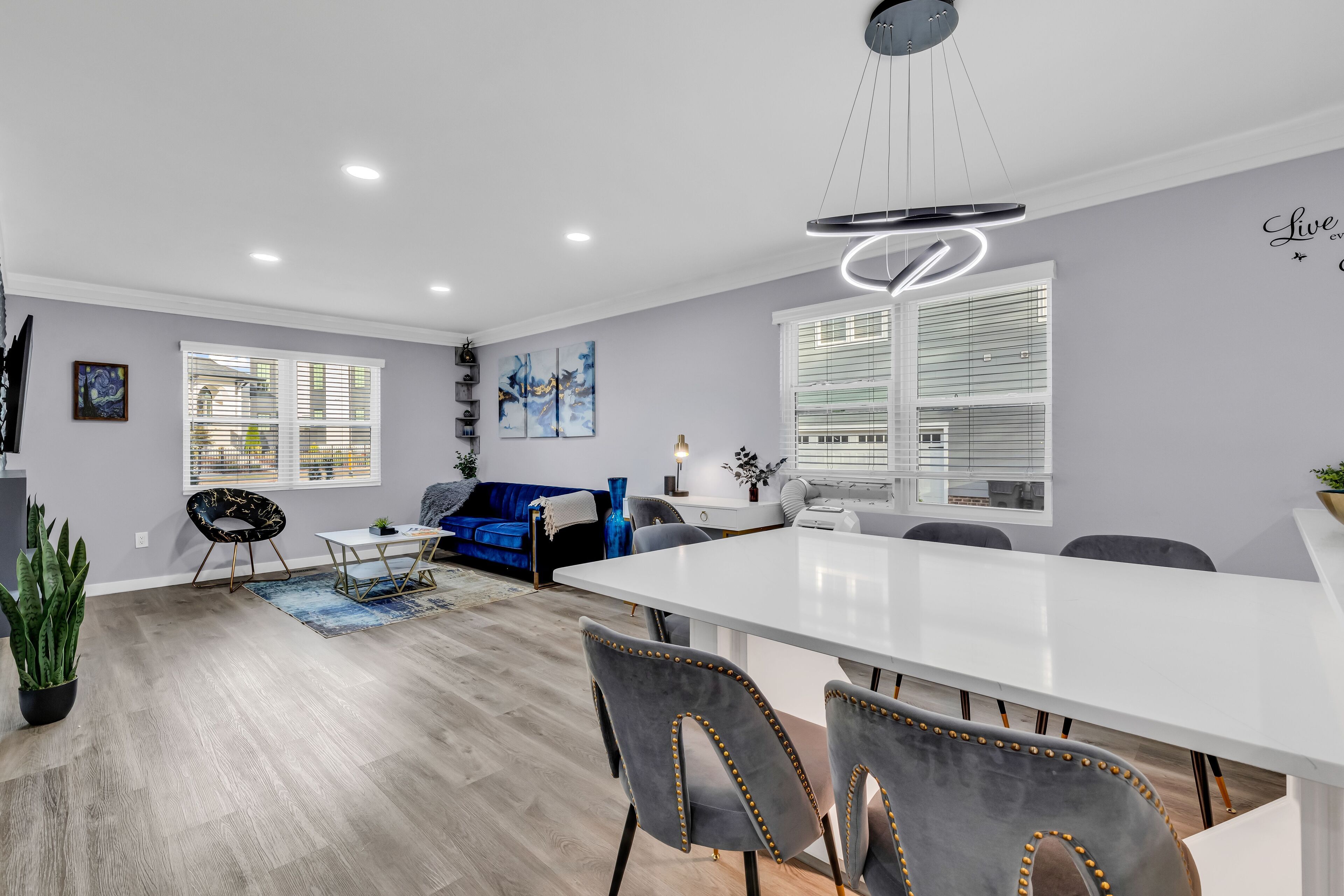 A modern living room with a dining area, featuring a large white table, grey upholstered chairs, and a blue sofa.