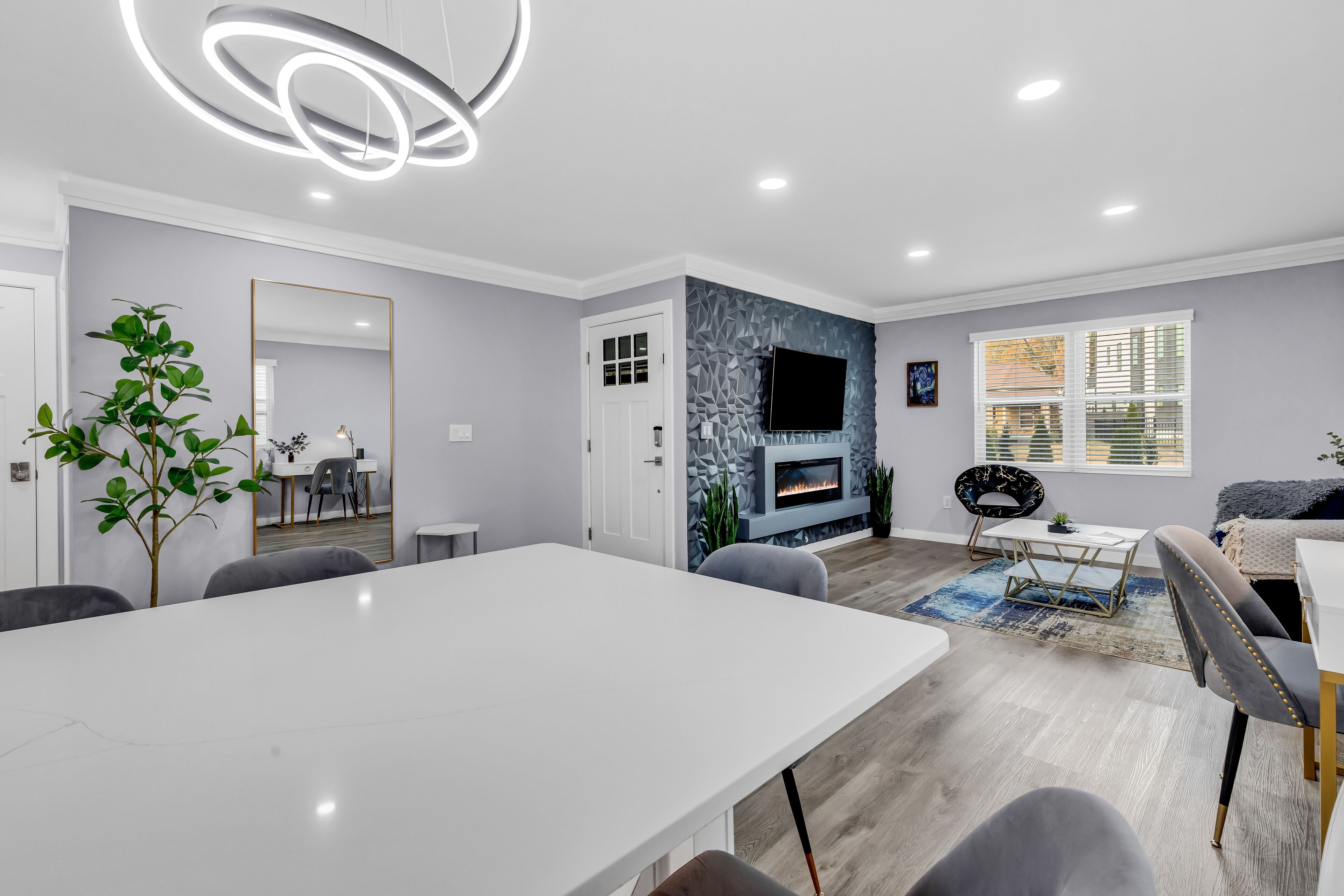 Een moderne woonkamer met een witte eettafel, grijze stoelen, een open haard en een aan de muur bevestigde televisie.