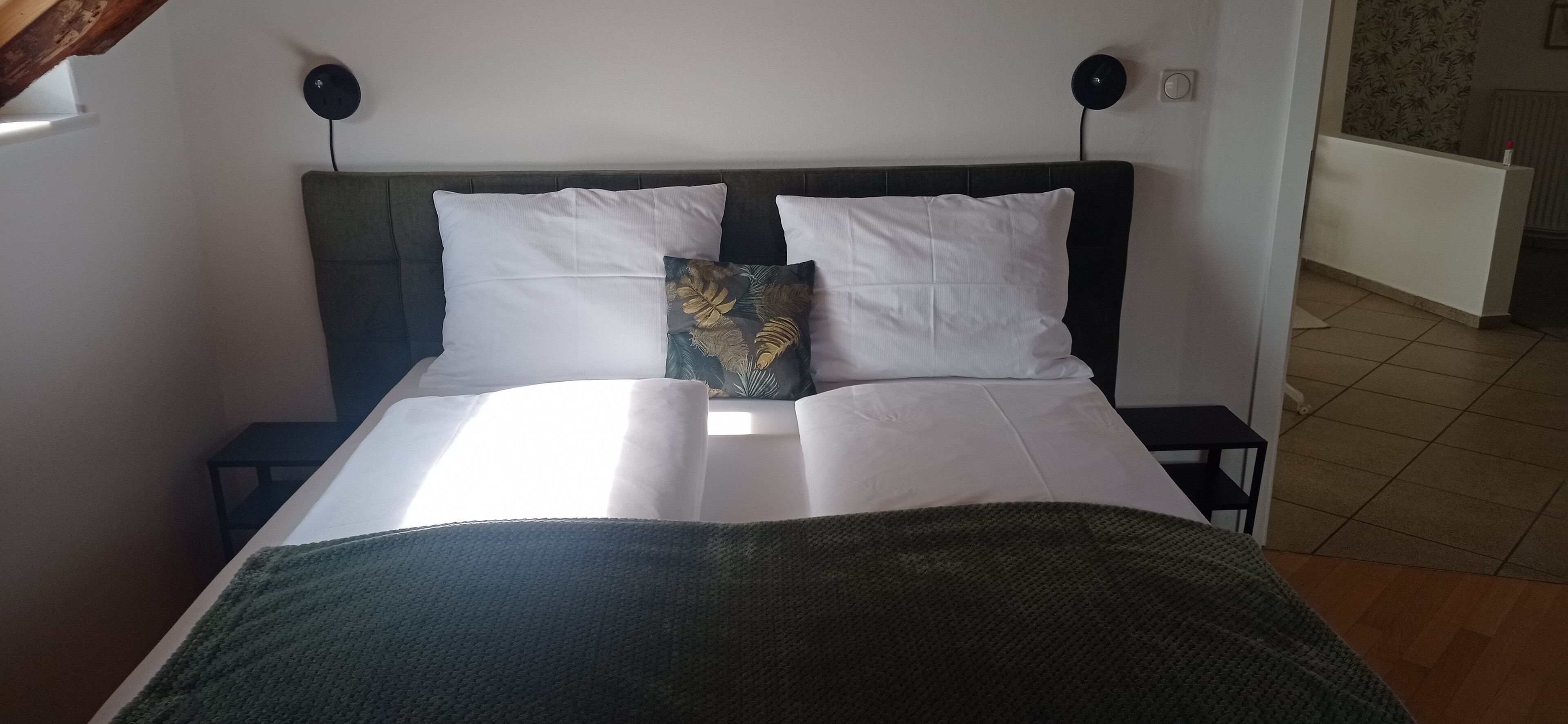 Una cama bien hecha con sábanas blancas y un plaid verde, dos almohadas blancas y una almohada decorativa con estampado de hojas. Hay dos mesitas de noche con lámparas negras.