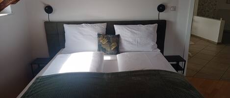 Una cama bien hecha con sábanas blancas y un plaid verde, dos almohadas blancas y una almohada decorativa con estampado de hojas. Hay dos mesitas de noche con lámparas negras.