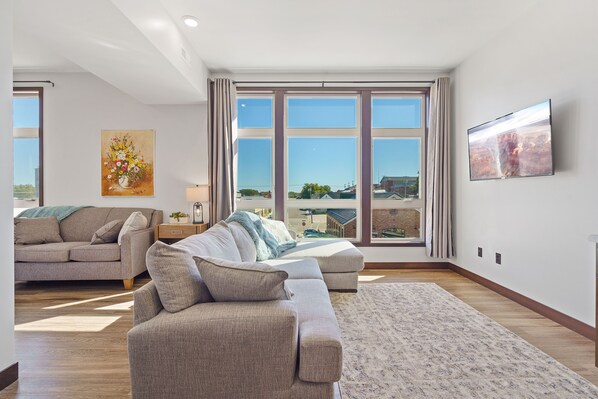 A spacious living room with a large window, a flat-screen TV, and a painting on the wall.