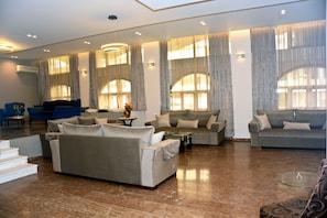 A spacious living area with multiple sofas and a coffee table, large windows with curtains, and a marble floor. - Hotel Marily (Pyrgos)