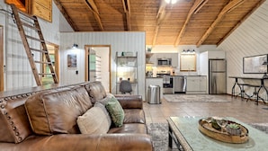 A spacious living area with a brown leather couch, a wooden coffee table with wine glasses, and a kitchen in the background.