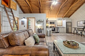 A spacious living area with a brown leather couch, a wooden coffee table with wine glasses, and a kitchen in the background.