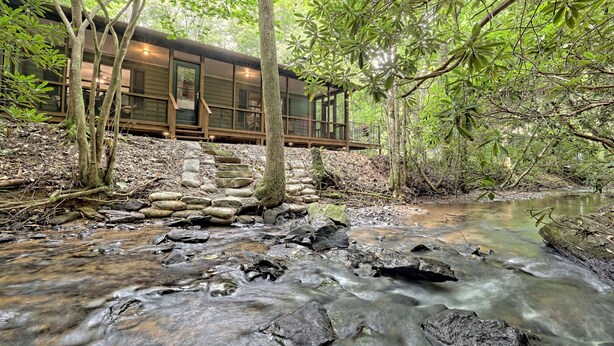 A wooden cabin with a stone staircase leading to a deck, surrounded by dense forest and a flowing stream.