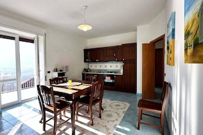 Uma área de refeições com mesa e cadeiras, uma cozinha com armários de madeira e vista através de uma porta de vidro.