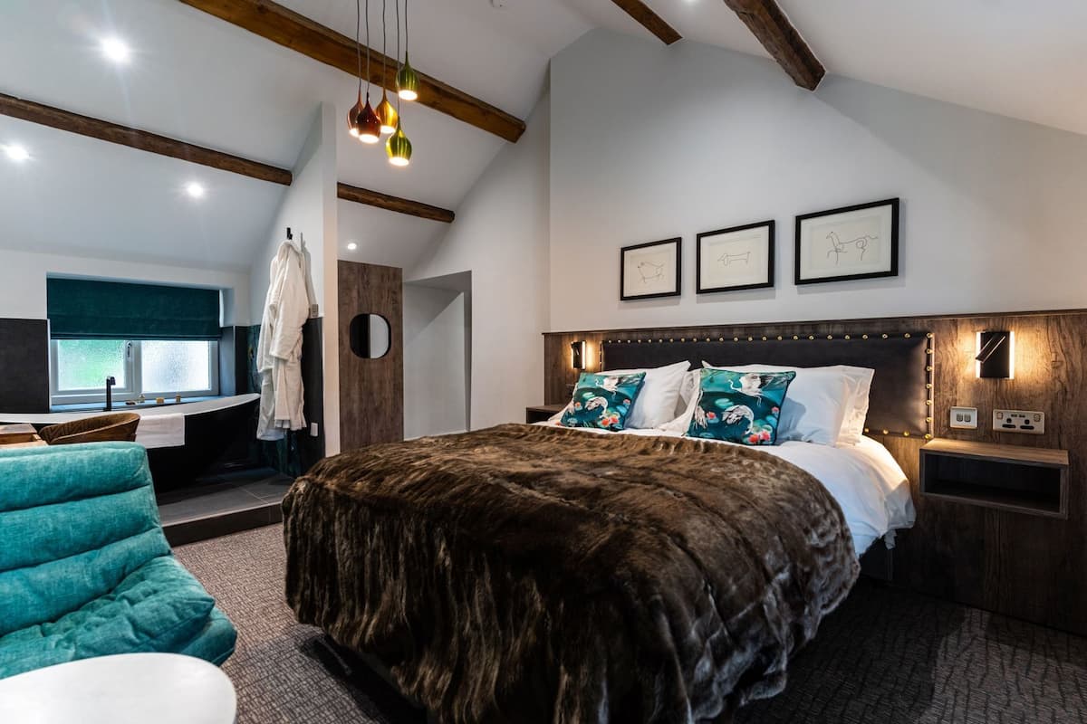 A modern hotel room with a large bed, a sitting area with a teal chair, a vanity with a mirror, and a wooden ceiling.