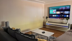A modern living room with a flat-screen TV mounted on a white wall, a grey sofa, a small coffee table, and a decorative plant. - A Home A Way from your Home with 3-bedroom & 2 bathroom with free parking  (Portland)