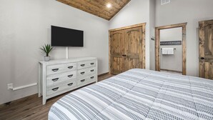 A bedroom with a wooden ceiling, a white dresser with drawers, a flat-screen TV, a large bed with a patterned bedspread, and wooden doors. - Private HotTub - Pool Front - Boat Slip Included (Branson)
