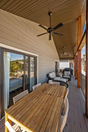 Een houten terras met een tafel, stoelen, een plafondventilator en uitzicht op een huis door een schuifdeur.