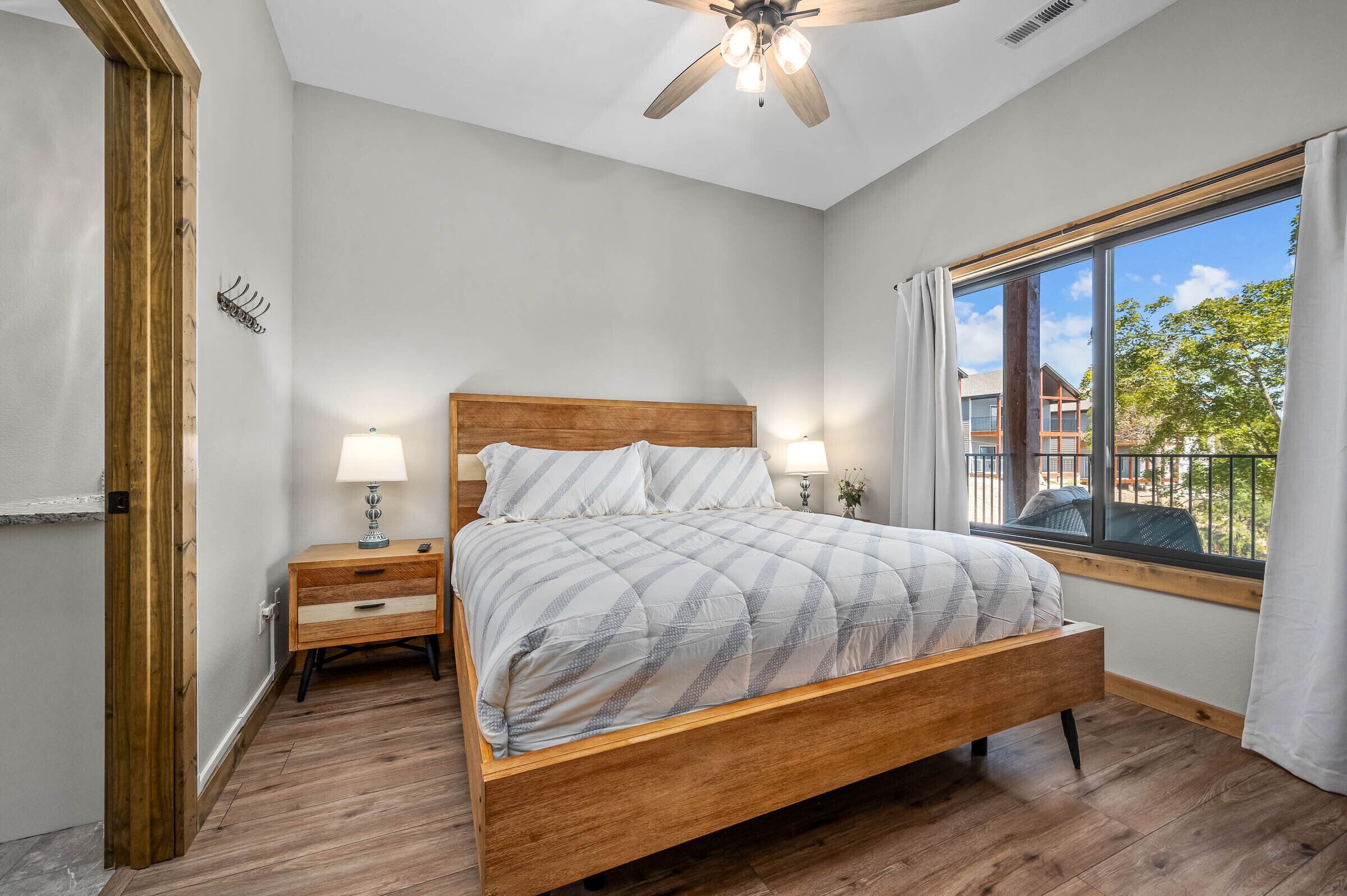 A bedroom with a wooden bed, two bedside tables with lamps, a ceiling fan, and a large window with a view of buildings and trees.