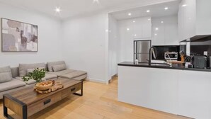 Een moderne woonkamer met een bank, salontafel en kunstwerk. In de achtergrond is een keuken te zien met witte kasten en apparatuur.
