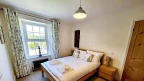 A bedroom with a wooden bed, white bedding, a bedside table with a lamp, a radiator, and a window with curtains. - Charming cottage with character features (Cornwall)