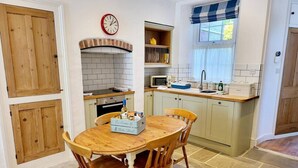 A kitchen with a wooden table, chairs, a clock, a microwave, and a window with a blue and white checkered curtain. - Charming cottage with character features (Cornwall)
