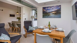A dining area with a wooden table set for four, surrounded by grey chairs. A painting of a landscape is on the wall above the table. - Modern, luxury bungalow - perfect for families or friends to relax! (Cornwall)