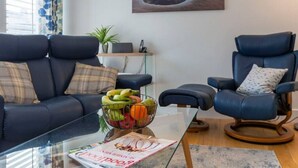 A modern living room with a sofa, two armchairs, a glass coffee table with a fruit bowl, and a magazine. - Modern, luxury bungalow - perfect for families or friends to relax! (Cornwall)