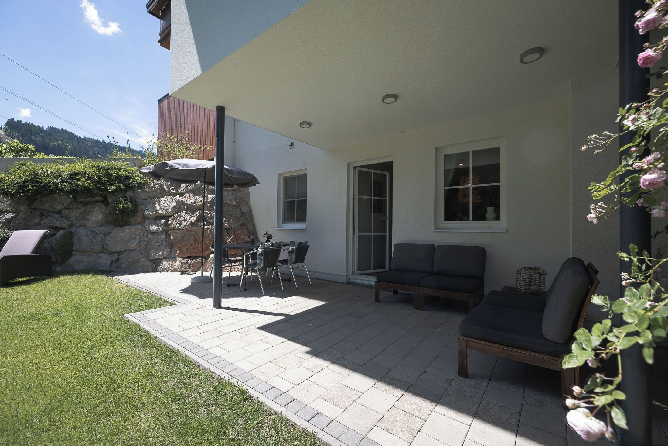 Eine Terrassenfläche mit einer Steinmauer, einem Tisch mit Stühlen, einer Sitzecke und einer Pergola.