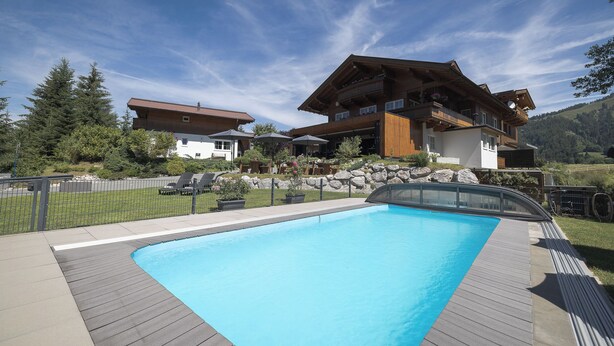 A pool area with a wooden deck, a mountain backdrop, and a chalet-style building with balconies. - Feriendorf-wallenburg / Enzian - Ferienwohnung für 2-4 Personen (Fieberbrunn)