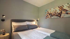 A neatly made bed with white and grey bedding, a bedside table with a lamp, and a wall-mounted painting. - Feriendorf-wallenburg / Enzian - Ferienwohnung für 2-4 Personen (Fieberbrunn)