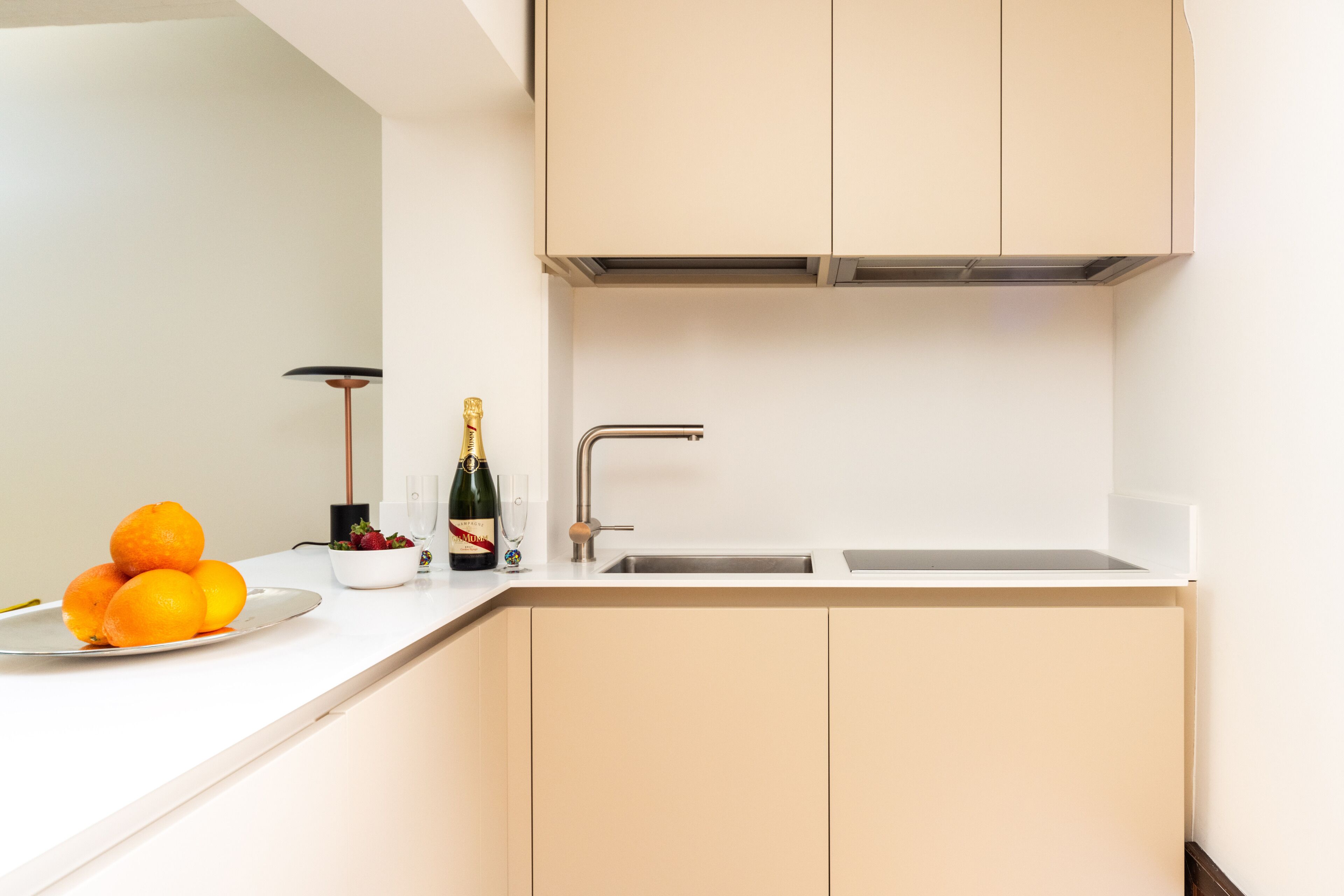 A modern kitchen with a white countertop, a sink, and cabinets. There are oranges on a plate, a bottle of champagne, and a bowl of fruit.
