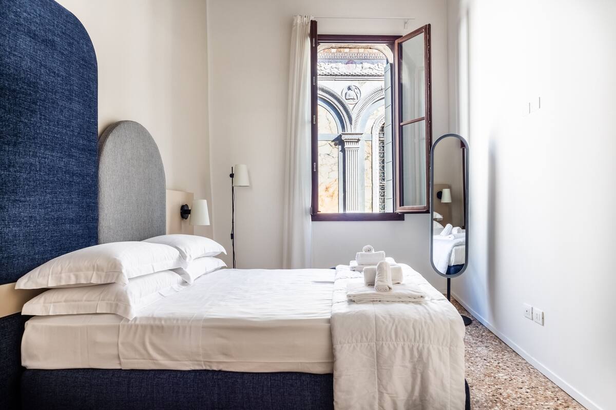 A neatly made bed with white linens, a large mirror, and a view of an ornate architectural detail through a window.