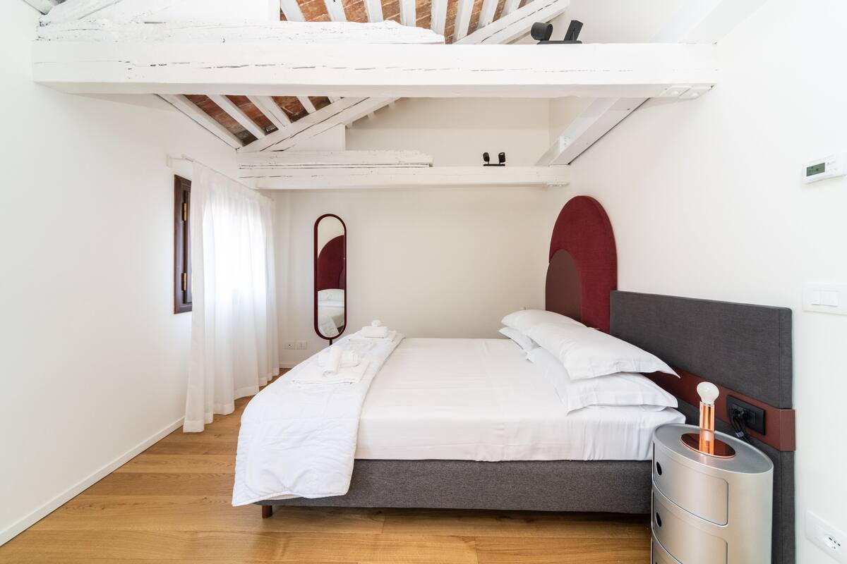 A bedroom with a bed, a bedside table, a mirror, and a window with curtains.