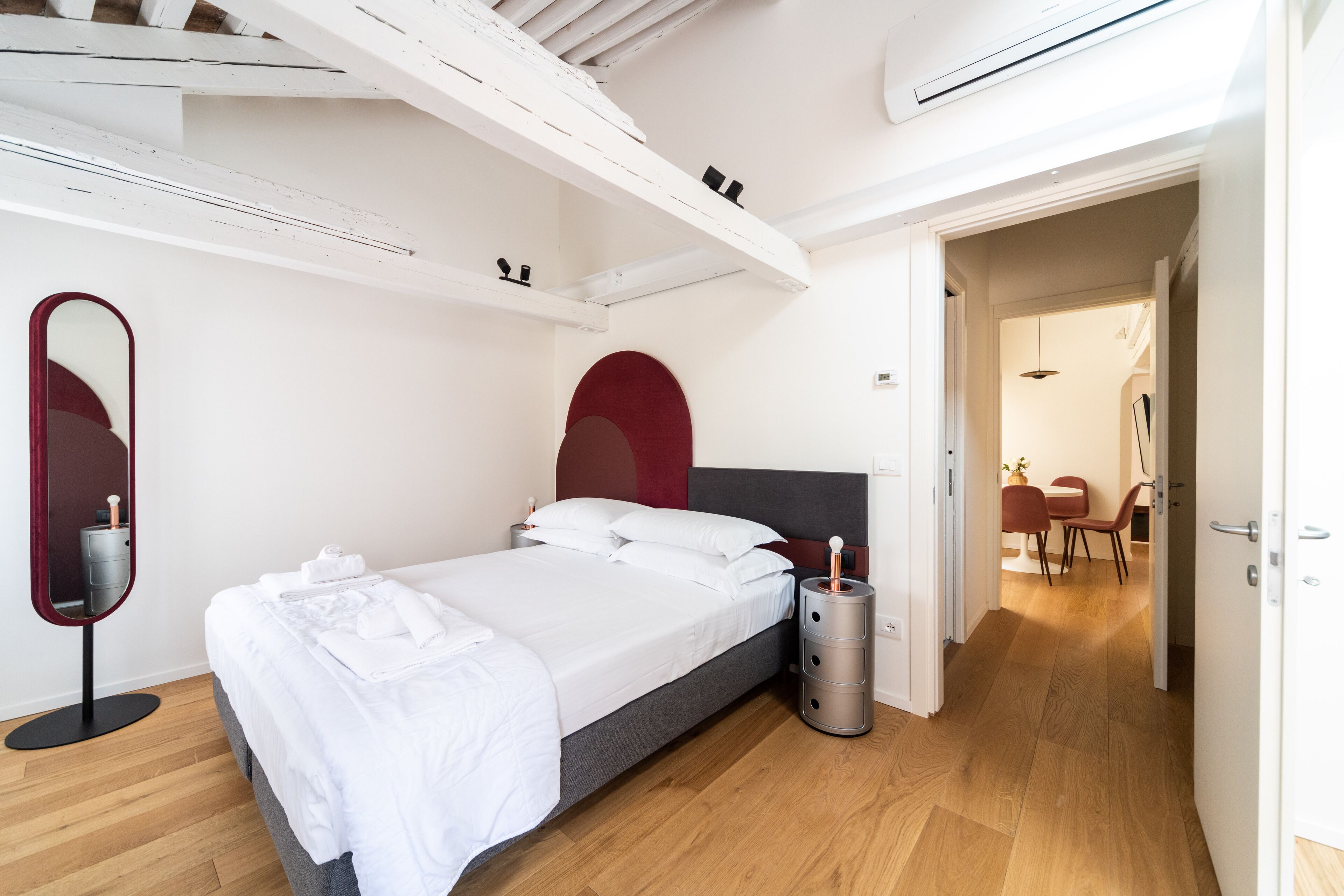 Un dormitorio moderno con cama, espejo junto a la cama, pared de acento roja y suelo de madera.