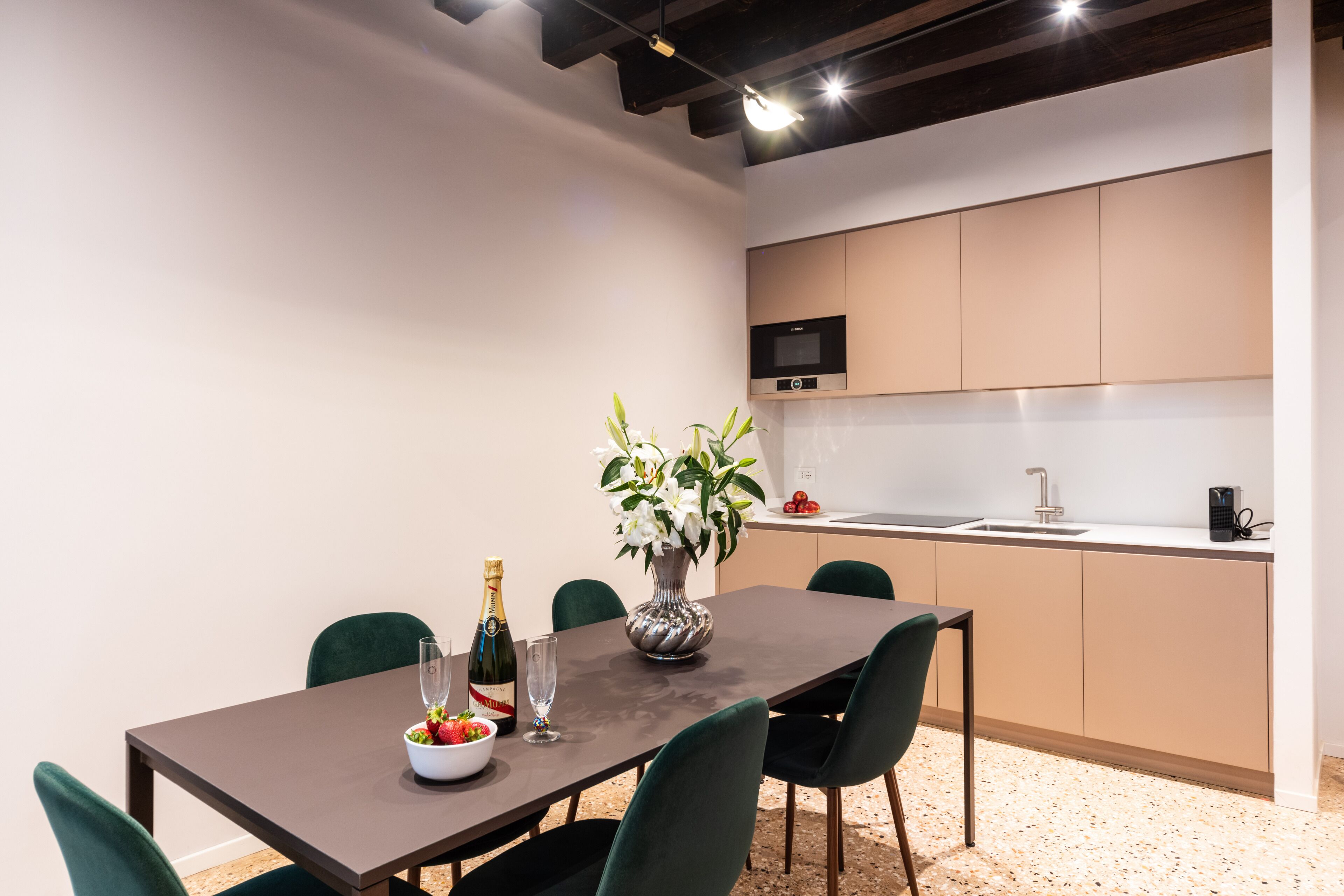 A modern dining area with a dark wooden table, green chairs, a vase with flowers, a bottle of champagne, and a bowl of fruit.