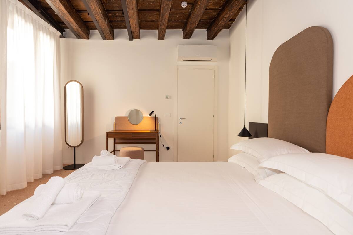 A hotel room with a large bed, a desk with a mirror, a chair, and a door.