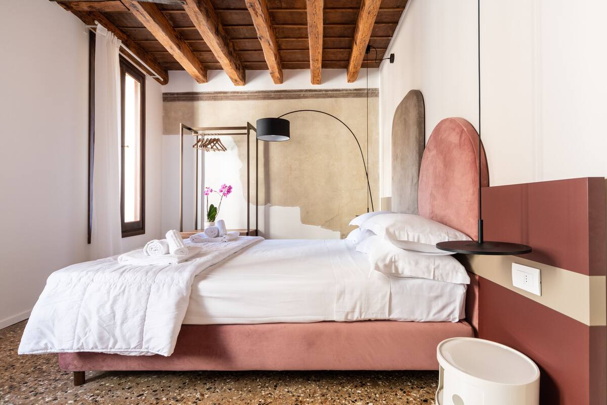 A bedroom with a bed, a bedside lamp, a potted plant, and a view of the outside.