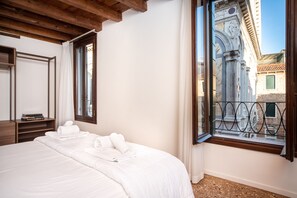 A hotel room with a large bed, a wooden ceiling, a window with a view of a building, and a small table with books.