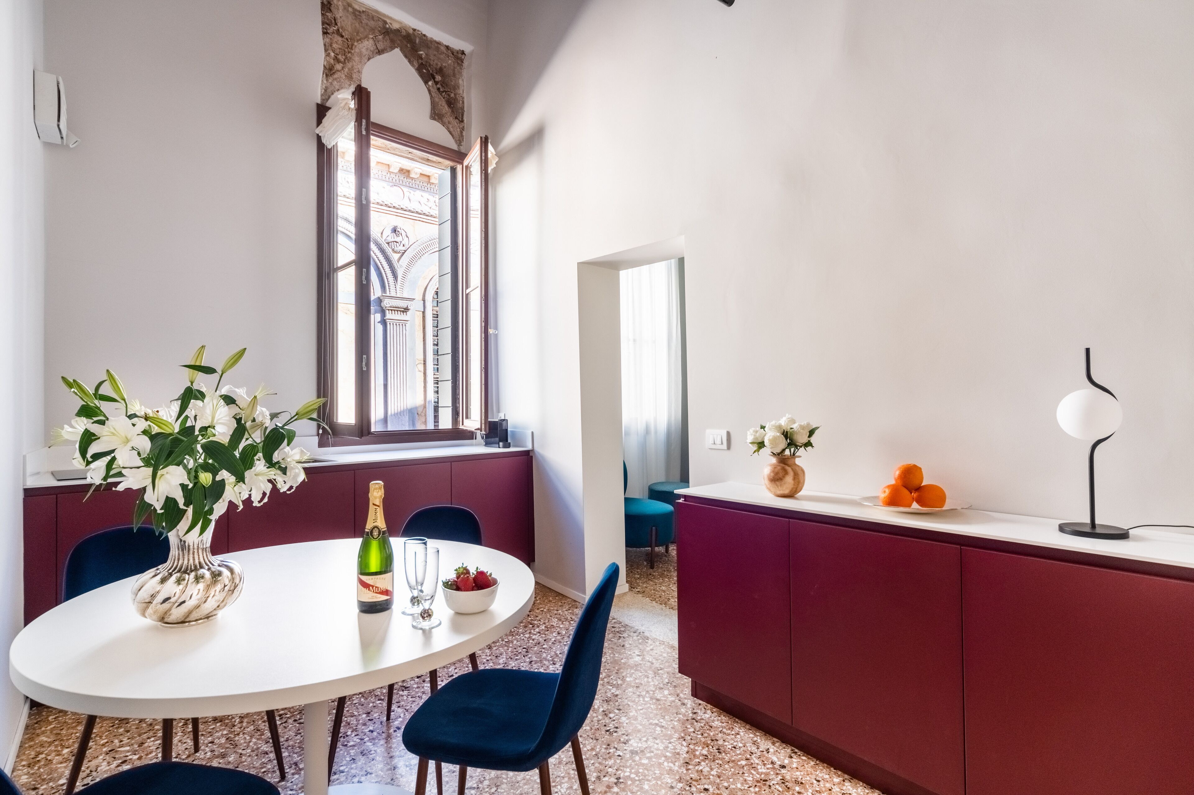A dining area with a white table, blue chairs, a vase of flowers, a bottle of champagne, and a bowl of fruit.