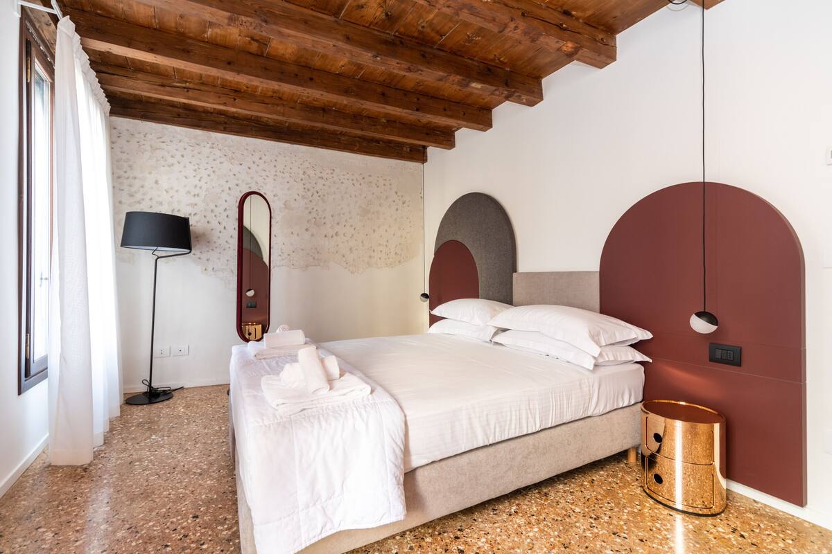 A bedroom with a bed, a bedside lamp, a round bedside table, and a wall-mounted mirror.