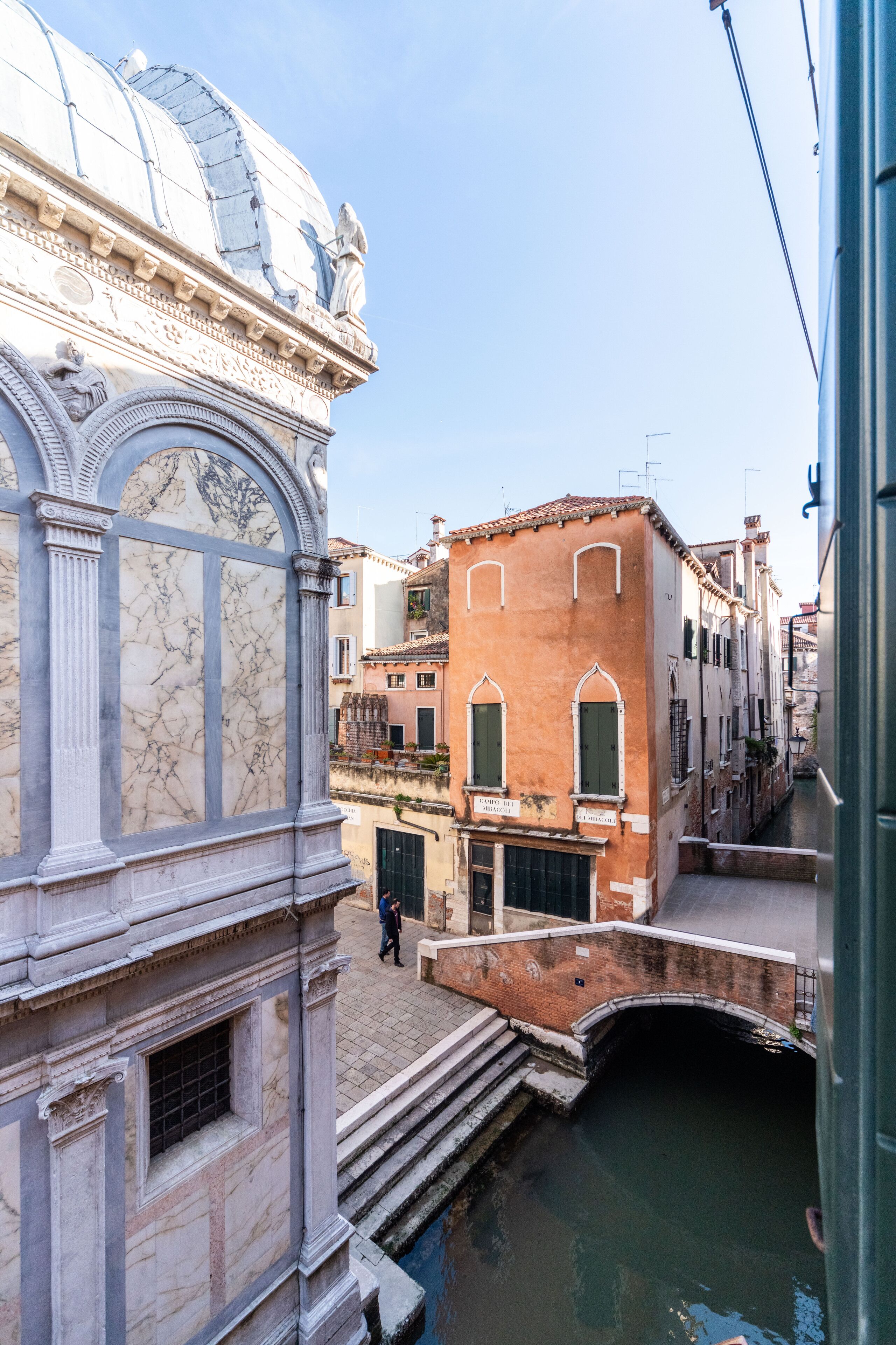 Un étroit canal vénitien bordé de bâtiments des deux côtés, dont une structure en marbre surmontée d’un dôme et un bâtiment rouge aux fenêtres en arc de cercle.