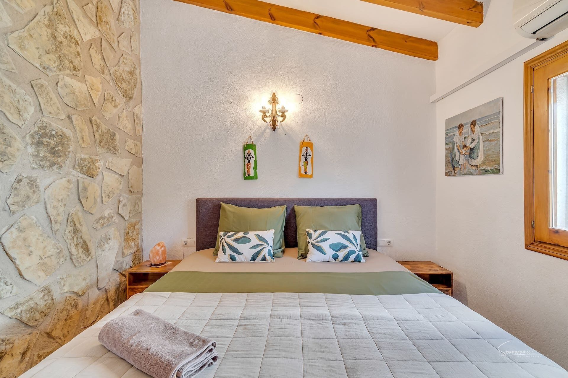 Une chambre avec un mur en pierre, un lit recouvert d’une courtepointe matelassée, deux oreillers, une table de chevet, un luminaire fixé au mur et un tableau accroché au mur.