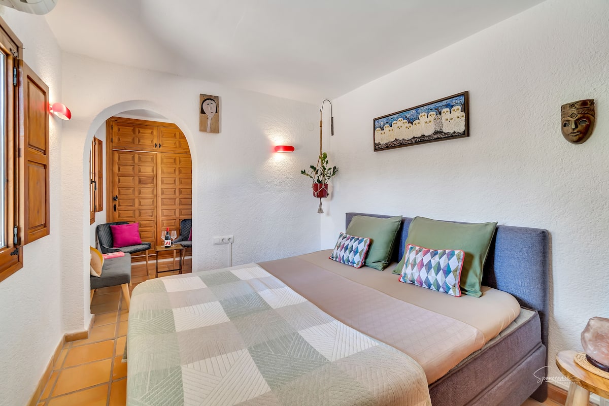 A bedroom with a bed, a desk, and a chair. There is a window with curtains, a door, and a wall-mounted light fixture.