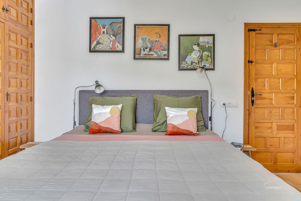 A neatly made bed with a grey and white duvet, two green pillows, and a red and white pillow. There are three framed artworks on the wall above the bed.