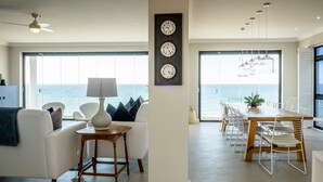 Een moderne woonkamer met uitzicht op zee, een eethoek met een houten tafel en stoelen, en een wand met drie klokken.