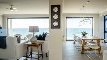A modern living room with a view of the sea, a dining area with a wooden table and chairs, and a wall with three clocks.