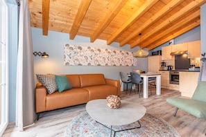 Ein gemütliches Wohnzimmer mit Holzbalkendecke, einem braunen Ledersofa, einem runden Couchtisch und einem Essbereich mit weißem Tisch und Stühlen.