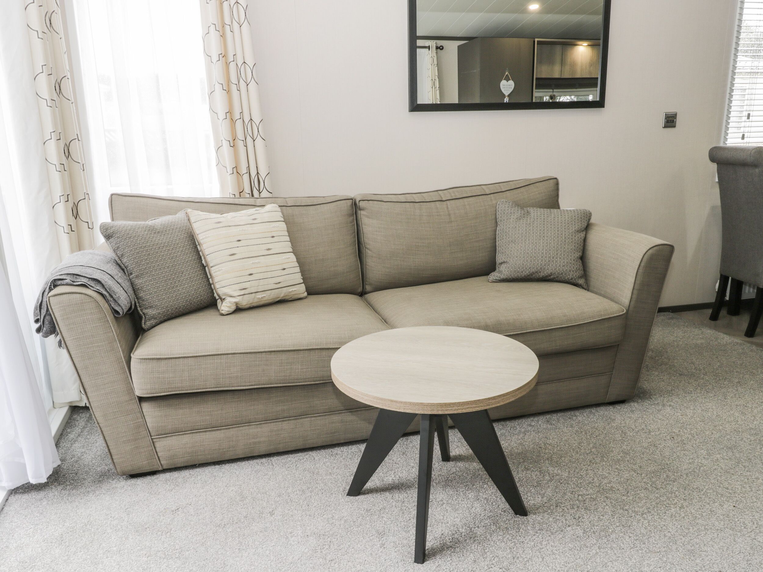 A beige sofa with patterned cushions, a round wooden table with black legs, and a mirror on the wall.