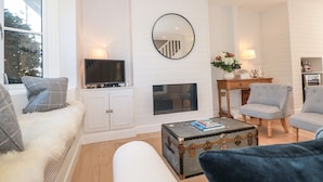 Ett modernt vardagsrum med en vit öppen spis, en tv, en stor spegel och en mysig sittgrupp med en bekväm soffa och fåtöljer.