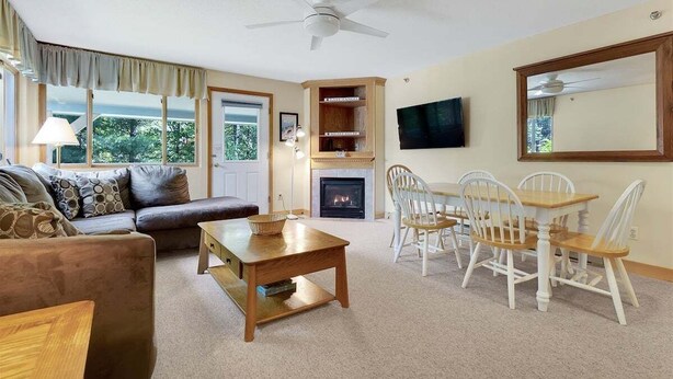 A living room with a sofa, a coffee table, a fireplace, a television, and a dining area with chairs. - Brookside I B-405 - Sunday River Slopeside Condominium (Newry)
