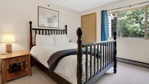 A bedroom with a bed, a nightstand, a ceiling fan, a window with curtains, and a door. - Brookside I B-405 - Sunday River Slopeside Condominium (Newry)