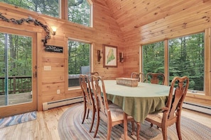 Interior de una cabaña de madera con una mesa de comedor, sillas, chimenea y ventanas grandes que dan a una vista del bosque.