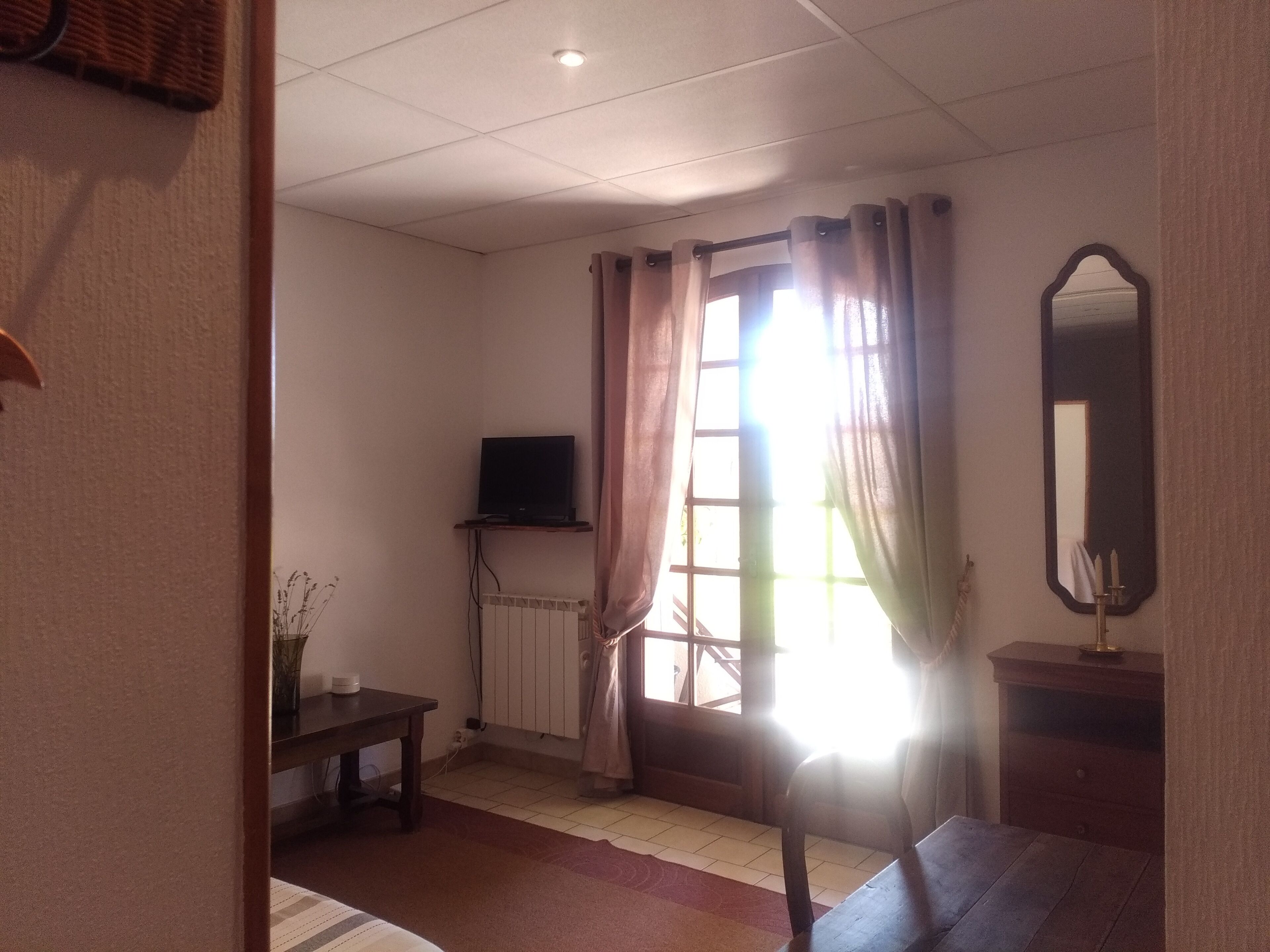 Een kamer met een televisie op een standaard, een radiator, een raam met gordijnen, een spiegel en een bed.